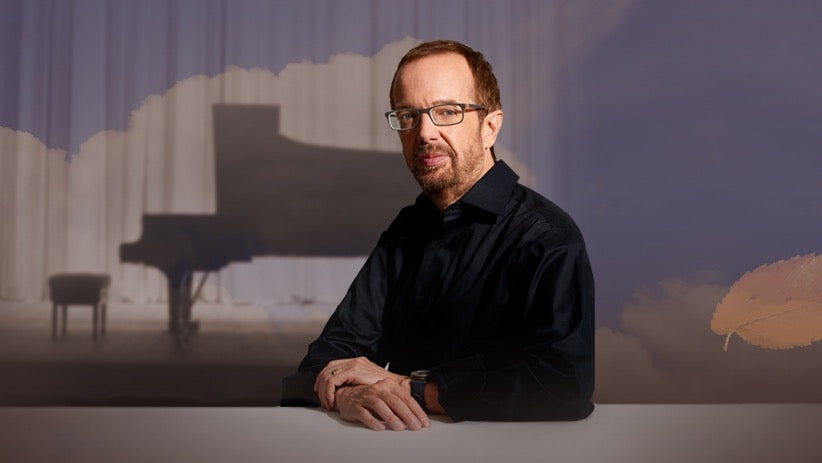 Steinway Artist, pianist and composer, Kevin Kern seated wearing black shirt and gray framed glasses. A concert grand Steinway piano silhouette is visible in the background.