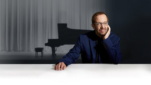 Steinway Artist and composer Kevin Kern, seated, wearing a blue jacket and glasses. His chin rests on his hand as he gazes towards the camera. There is a concert grand piano and piano bench on stage in the background.